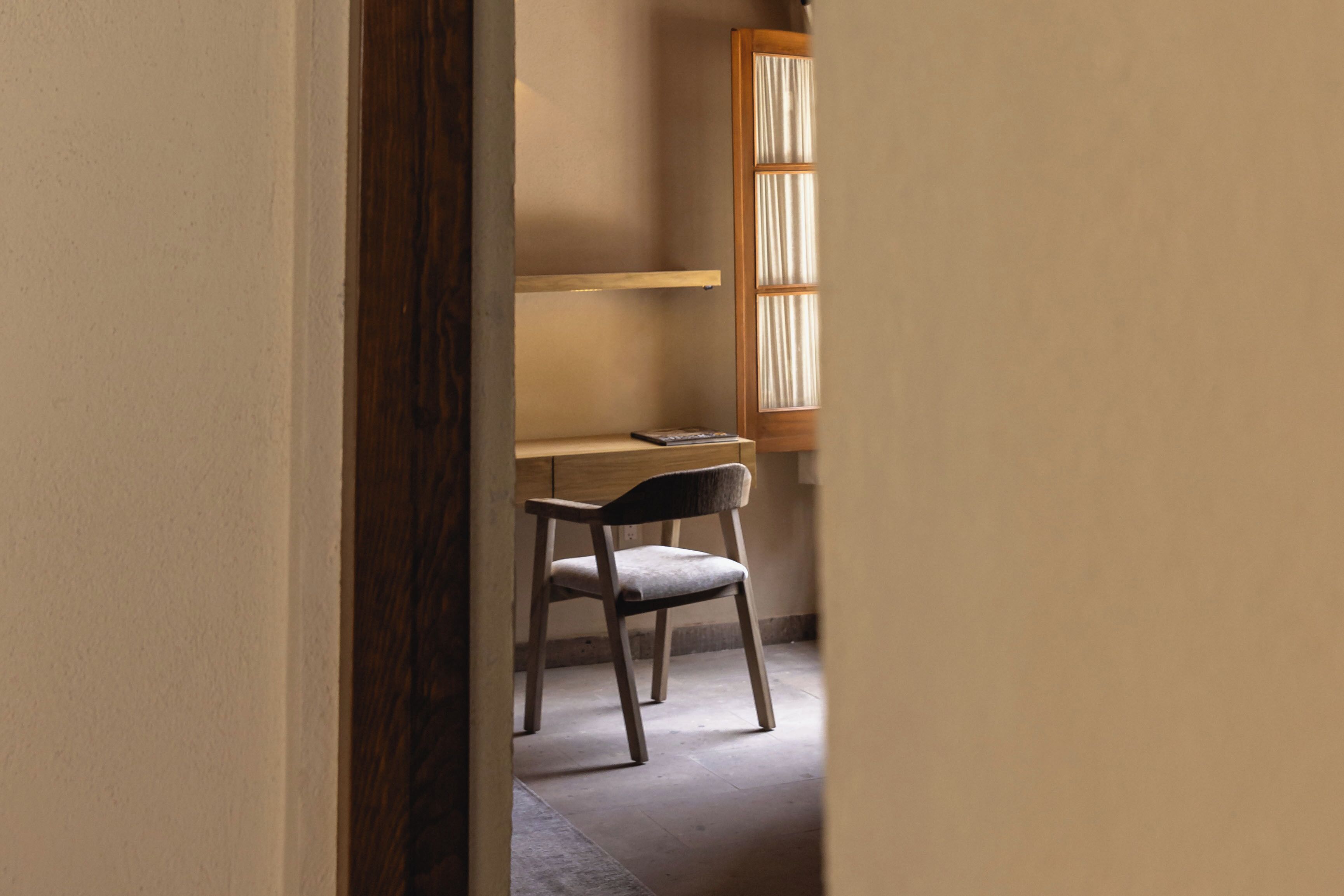 A narrow hallway with a wooden door frame, a desk with a chair, and a window with curtains.