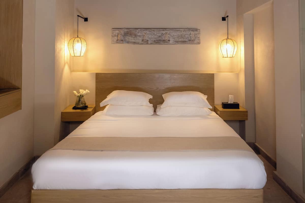 Una cama bien hecha con sábanas blancas, dos mesitas de noche con lámparas y una obra de arte rústica sobre la cama.