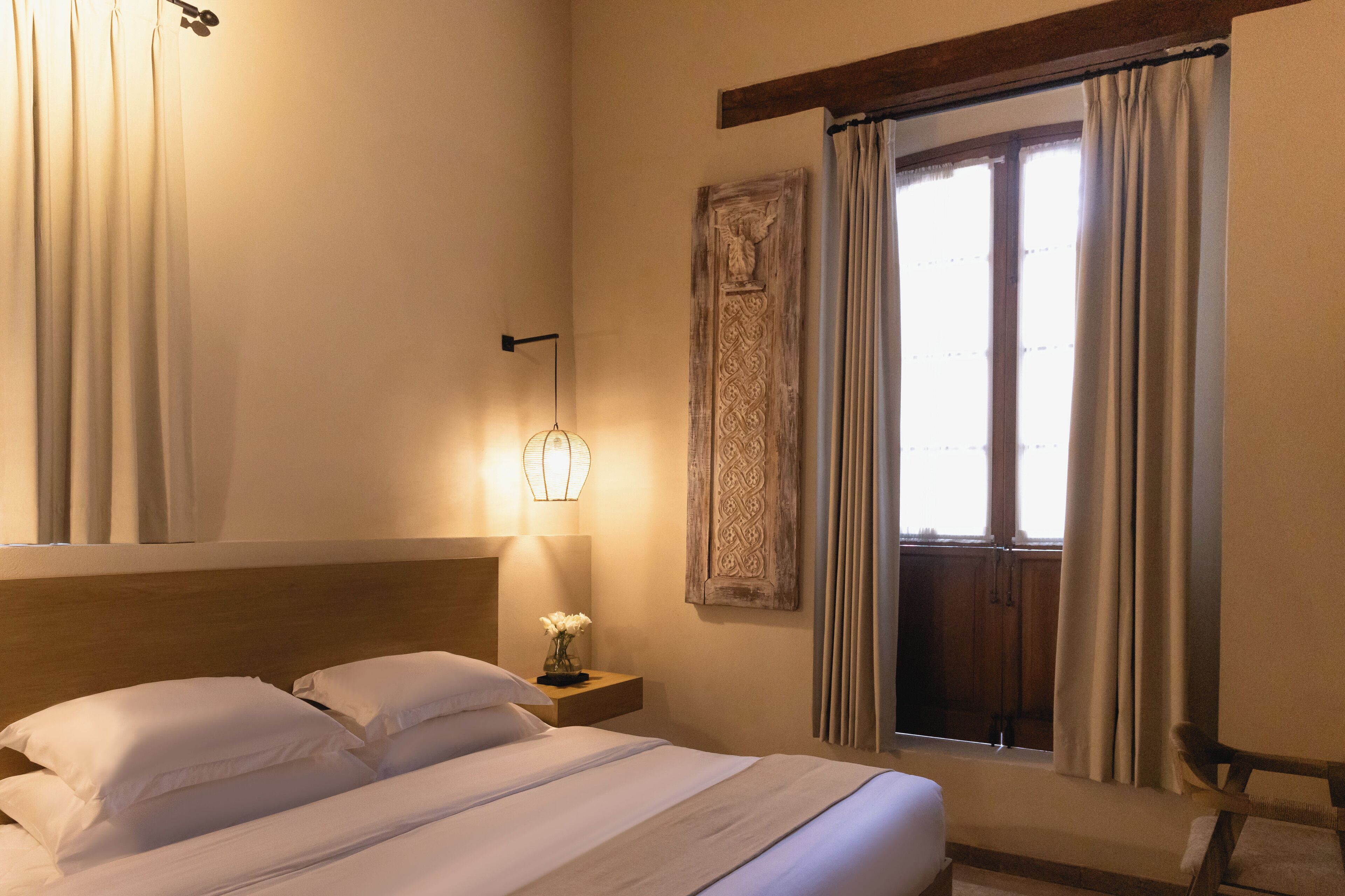 A neatly made bed with white linens, a bedside table with a lamp, and a window with curtains.