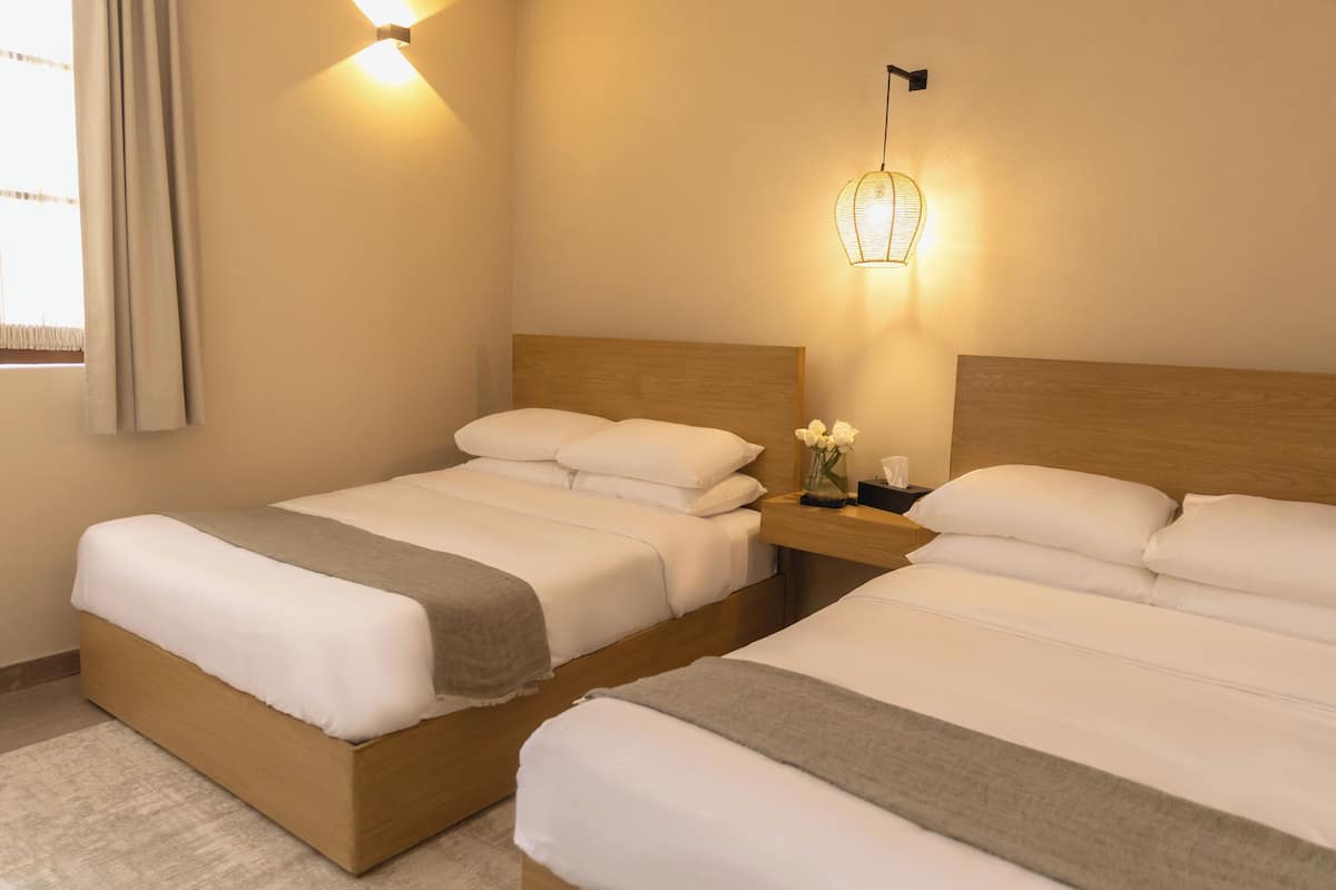 Habitación de hotel con dos camas, una mesita de noche, un aplique en la pared y un ventanal con cortinas.