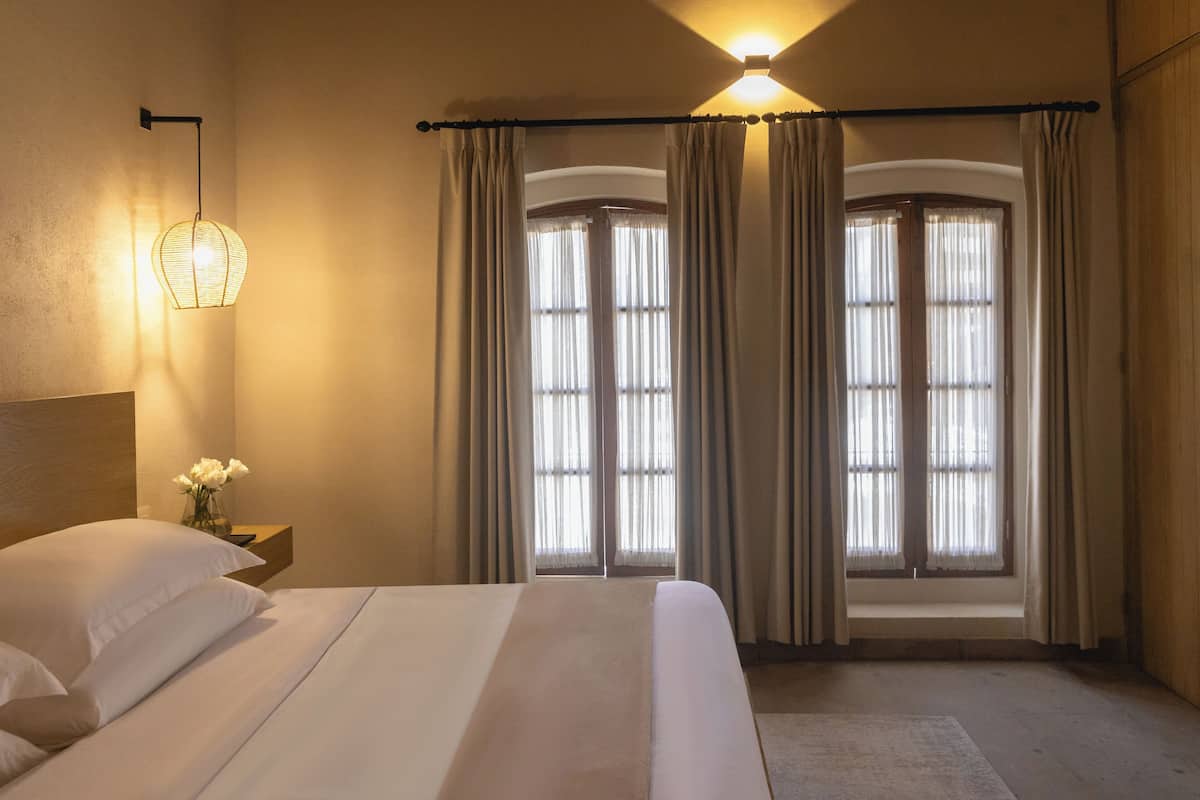 Una habitación de hotel con una cama grande, una mesita de noche con un jarrón de flores y una ventana con cortinas.