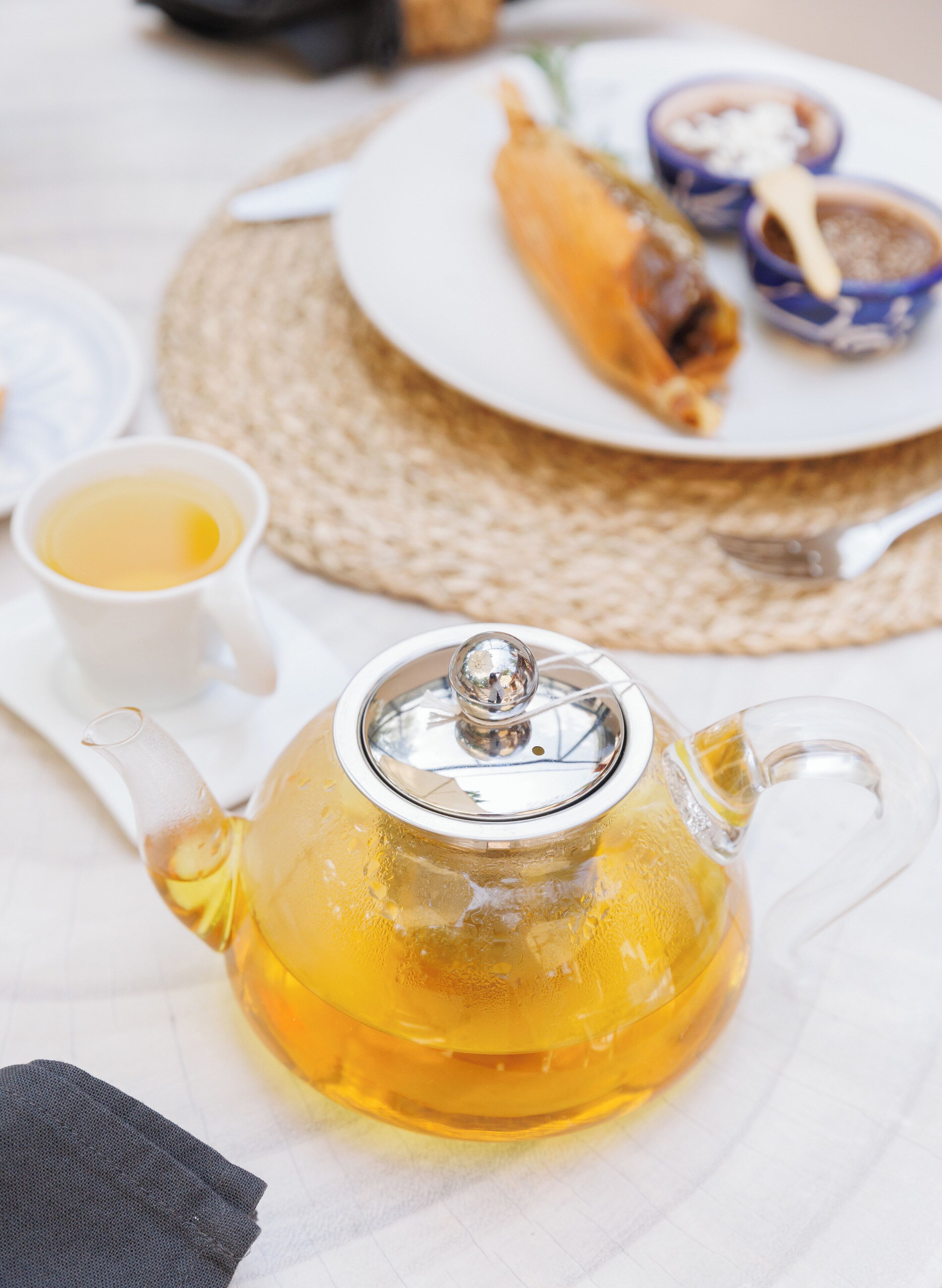 Una tetera de vidrio transparente con tapa, una taza blanca, un posavasos tejido y un plato con comida y cuencos azules.