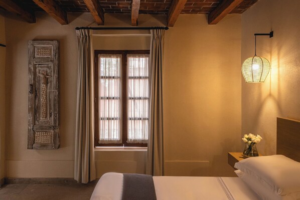 Un dormitorio con cama, mesita de noche con un florero, ventana con cortinas e iluminación en la pared.