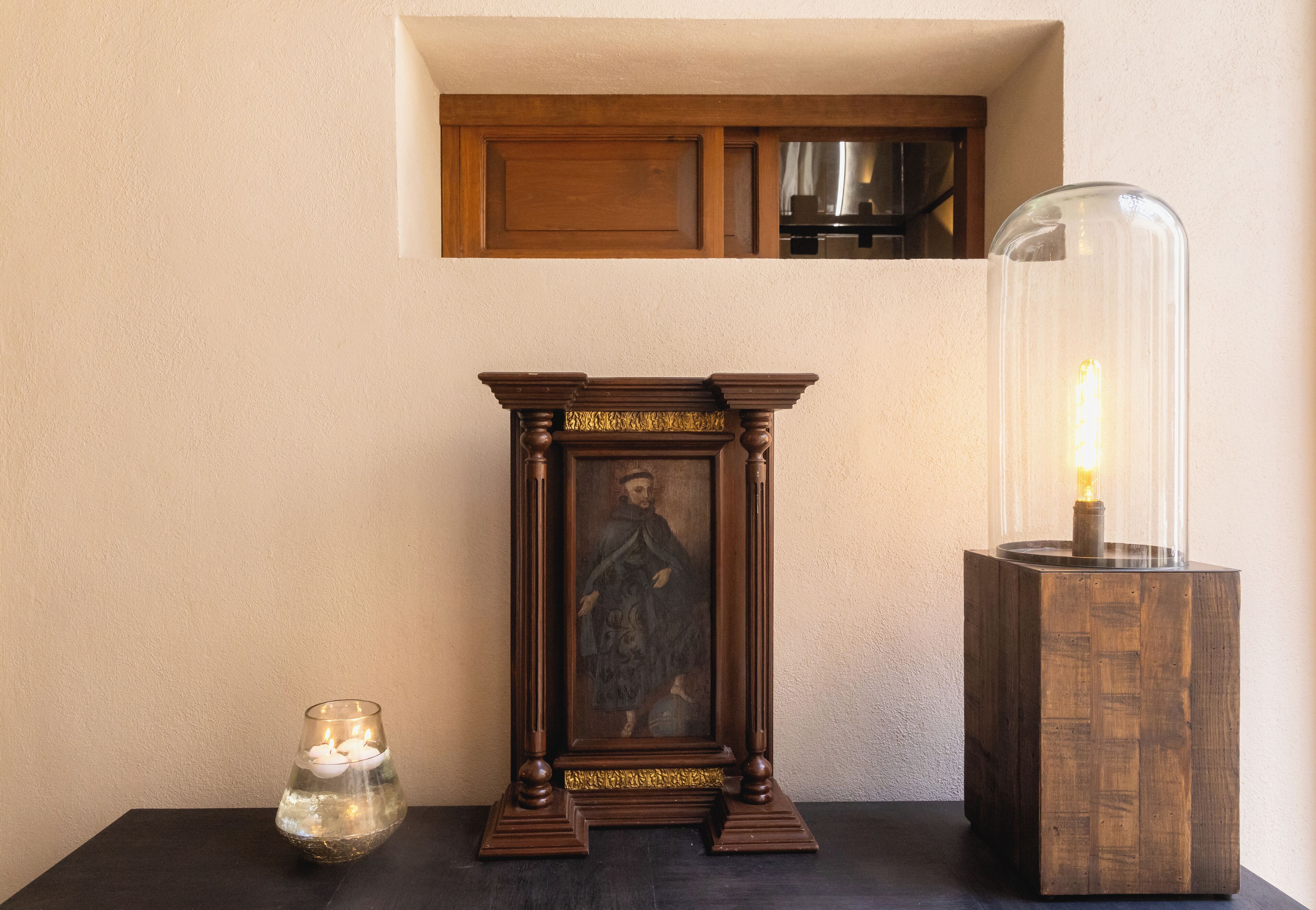 A framed religious painting, a candle in a glass holder, and a modern lamp on a dark table against a plain wall.