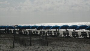 Ein Strand mit Reihen von blauen Sonnenschirmen und Liegestühlen, einer Holzbrüstung und dem Ozean im Hintergrund.