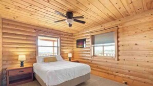 Ein Schlafzimmer in einer Blockhütte mit einem Bett, zwei Fenstern, einem Deckenventilator und einem Nachttisch mit einer Lampe.