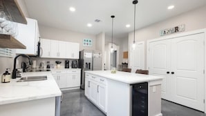 Una cocina moderna con armarios blancos, una isla central, electrodomésticos de acero inoxidable e iluminación colgante.