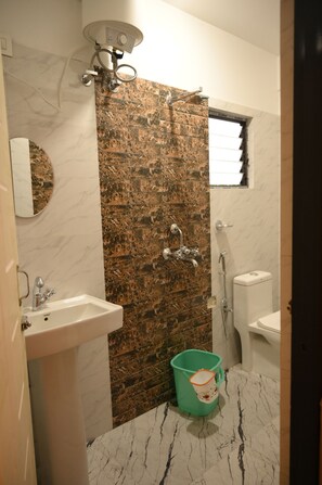 A bathroom with a marble sink, a green bucket, a shower with a wall-mounted faucet, and a window with blinds. - Bayaroo Stay Lamahatta (Darjeeling)