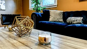 A living room with a wooden coffee table, a geometric decorative piece, a candle labeled "Vanilla," a dark blue sofa, and a window with a view of greenery. - Grand Lakeview Retreat Near Town • Game Rm • 4BR (Princeton)