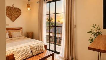 A bedroom with a bed, a bench, a bedside table, a wall-mounted light fixture, a large window with curtains, and a view of palm trees.