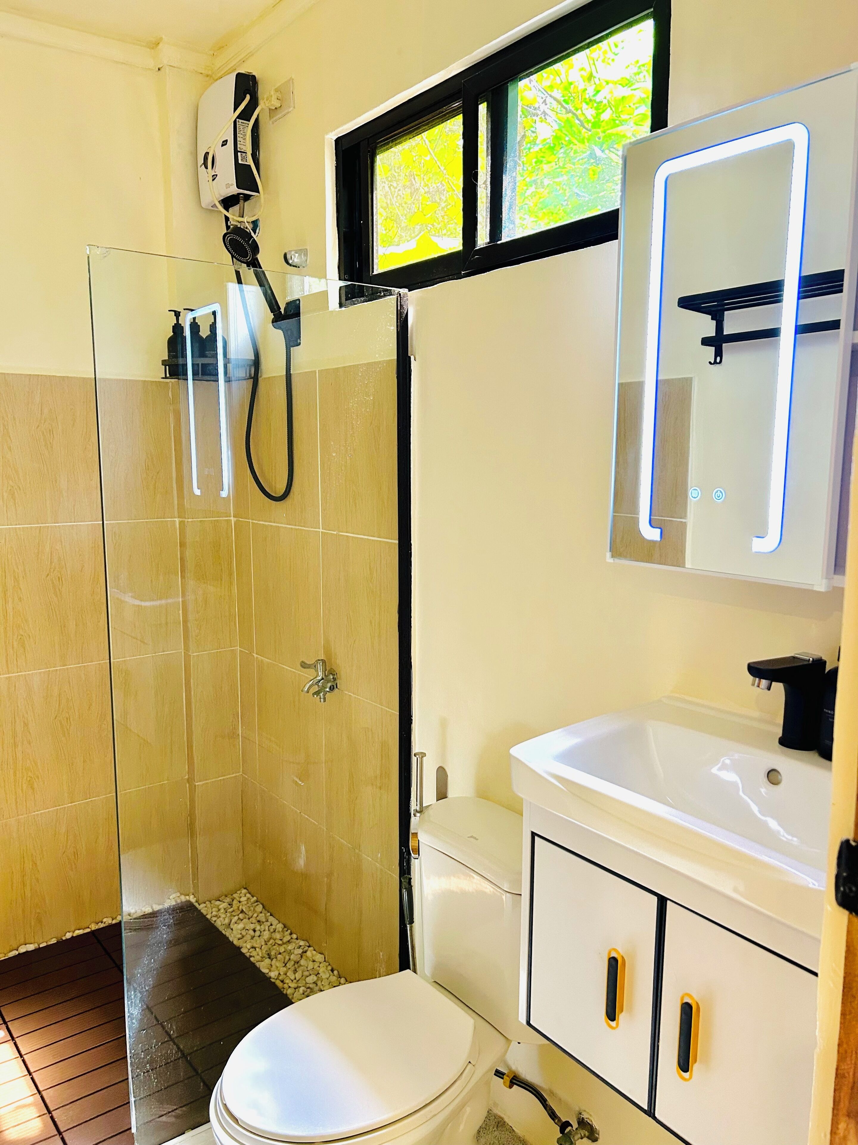 Un baño con cabina de ducha de vidrio, inodoro blanco, lavamanos con espejo y ventana con vistas a follaje.