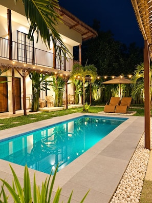 A swimming pool with lounge chairs and umbrellas, surrounded by tropical plants and a building with a balcony. - Lolas Villa El Nido (El Nido)