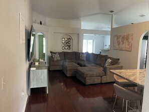 A modern living room with a sectional sofa, a dining area with a marble table, and a flat-screen TV mounted on the wall.