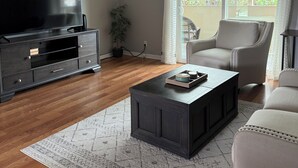 Un salon doté d’un téléviseur à écran plat, d’un meuble de rangement en bois foncé, d’un ventilateur de plafond, d’une table basse, d’un tapis à motifs et d’une grande fenêtre avec des rideaux.