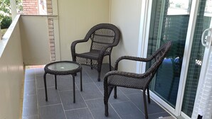 Un balcon doté d’un sol carrelé de couleur gris foncé, de meubles en osier et d’une porte vitrée.