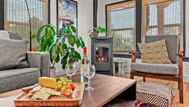A cozy living room with a fireplace, a sofa, a coffee table with snacks, and a large window with blinds.