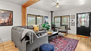 A modern living room with a gray sofa, a coffee table, and a patterned rug. There is a fireplace, a plant, and a window with a view of trees.