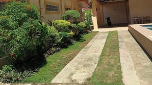 Zona exterior coberta amb un camí de formigó que condueix a l'entrada d'un edifici, envoltada de vegetació cuidada i un cel blau clar.