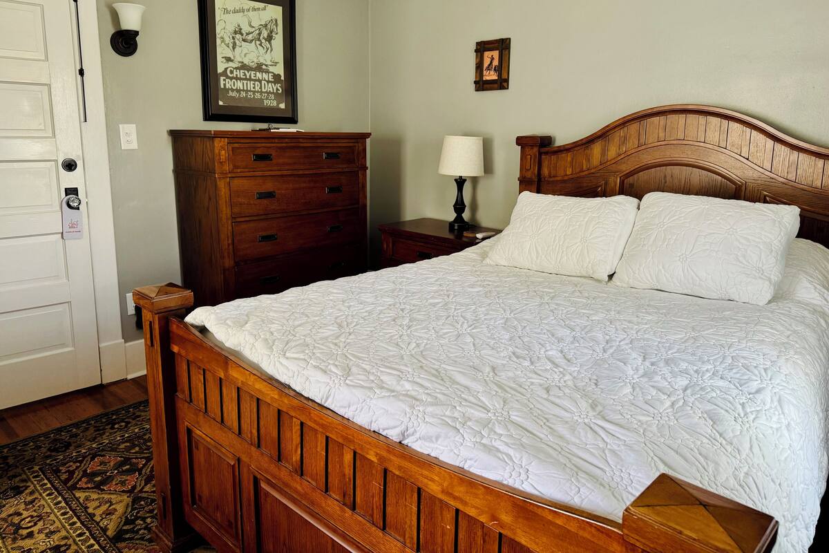 A bedroom with a wooden bed, a nightstand, a dresser, a framed picture on the wall, and a door with a lock.