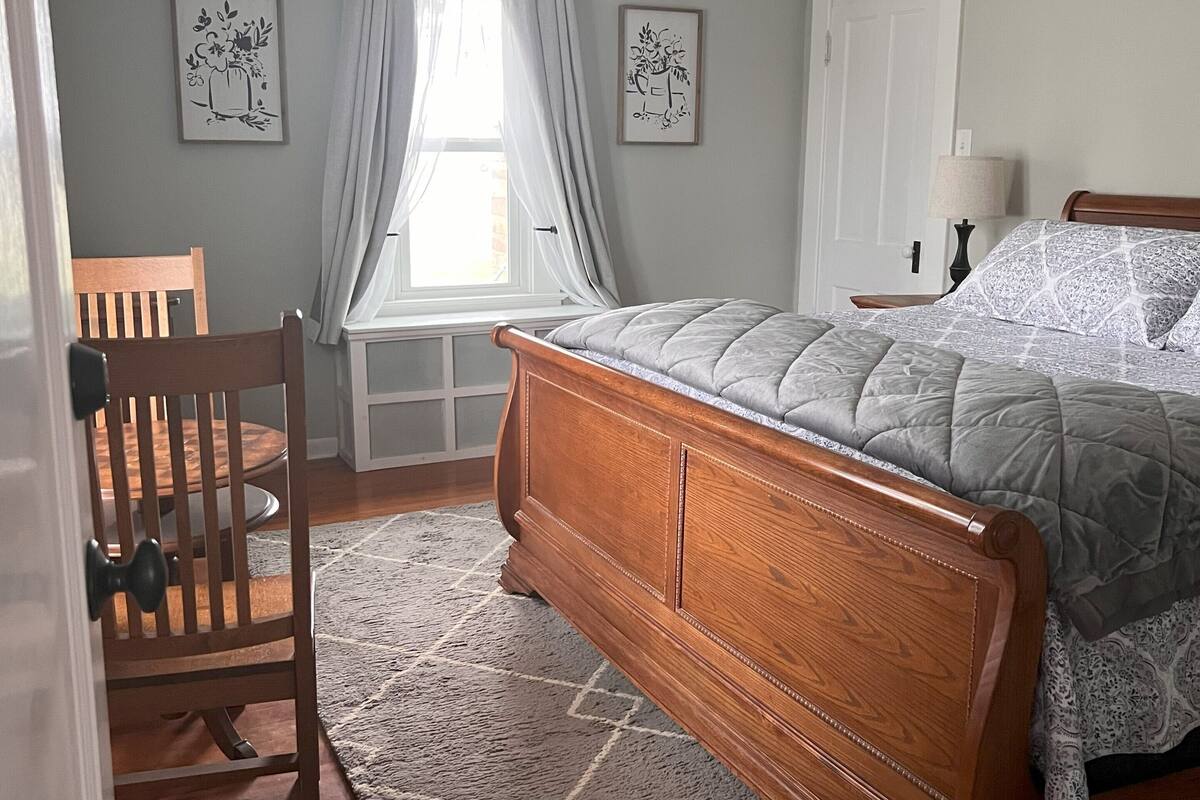 A bedroom with a wooden bed, a chair, a desk, and a window with curtains.