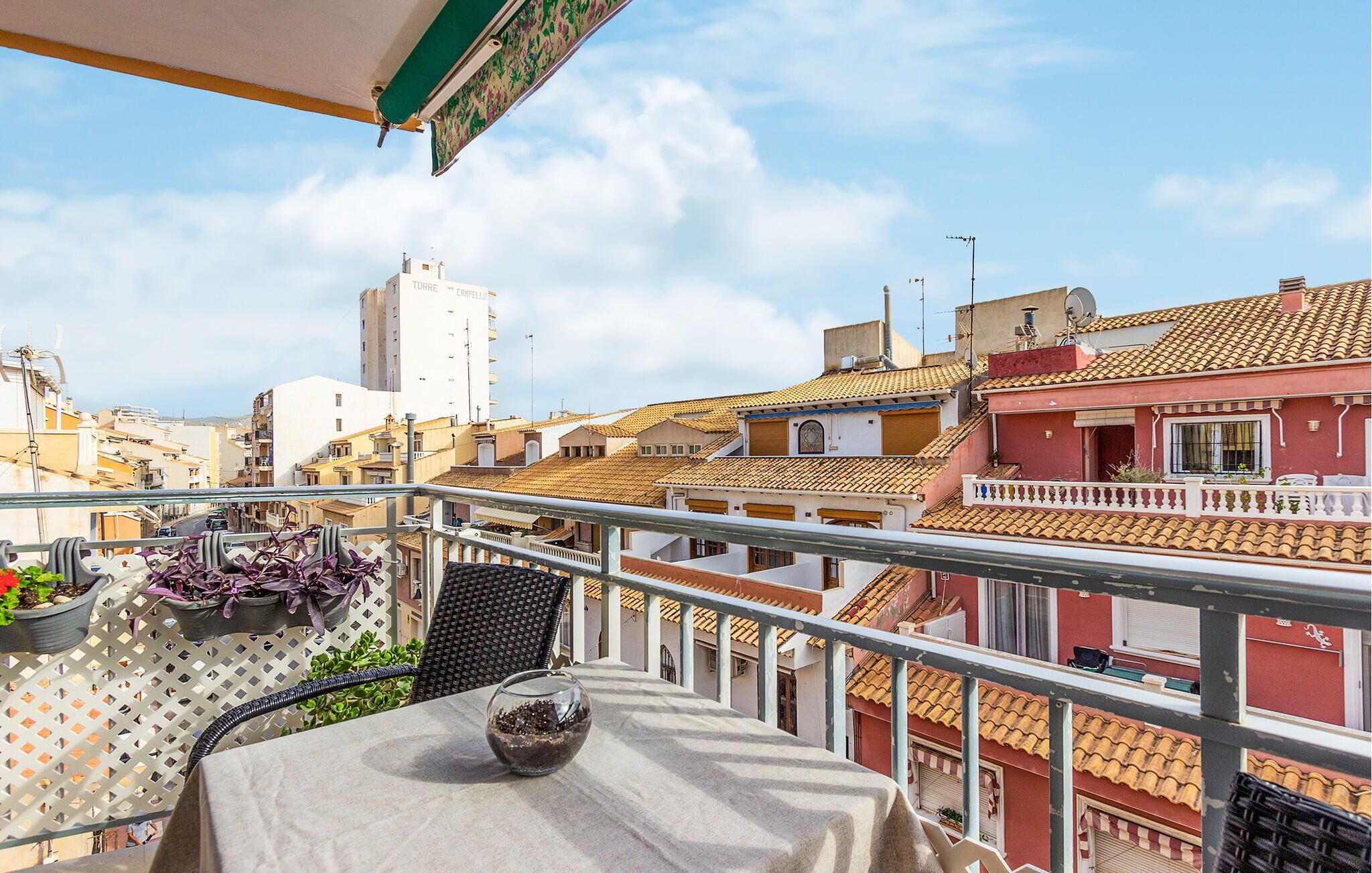 Un balcón con una mesa, plantas en macetas y vistas a un paisaje urbano con edificios de tejas de terracota.