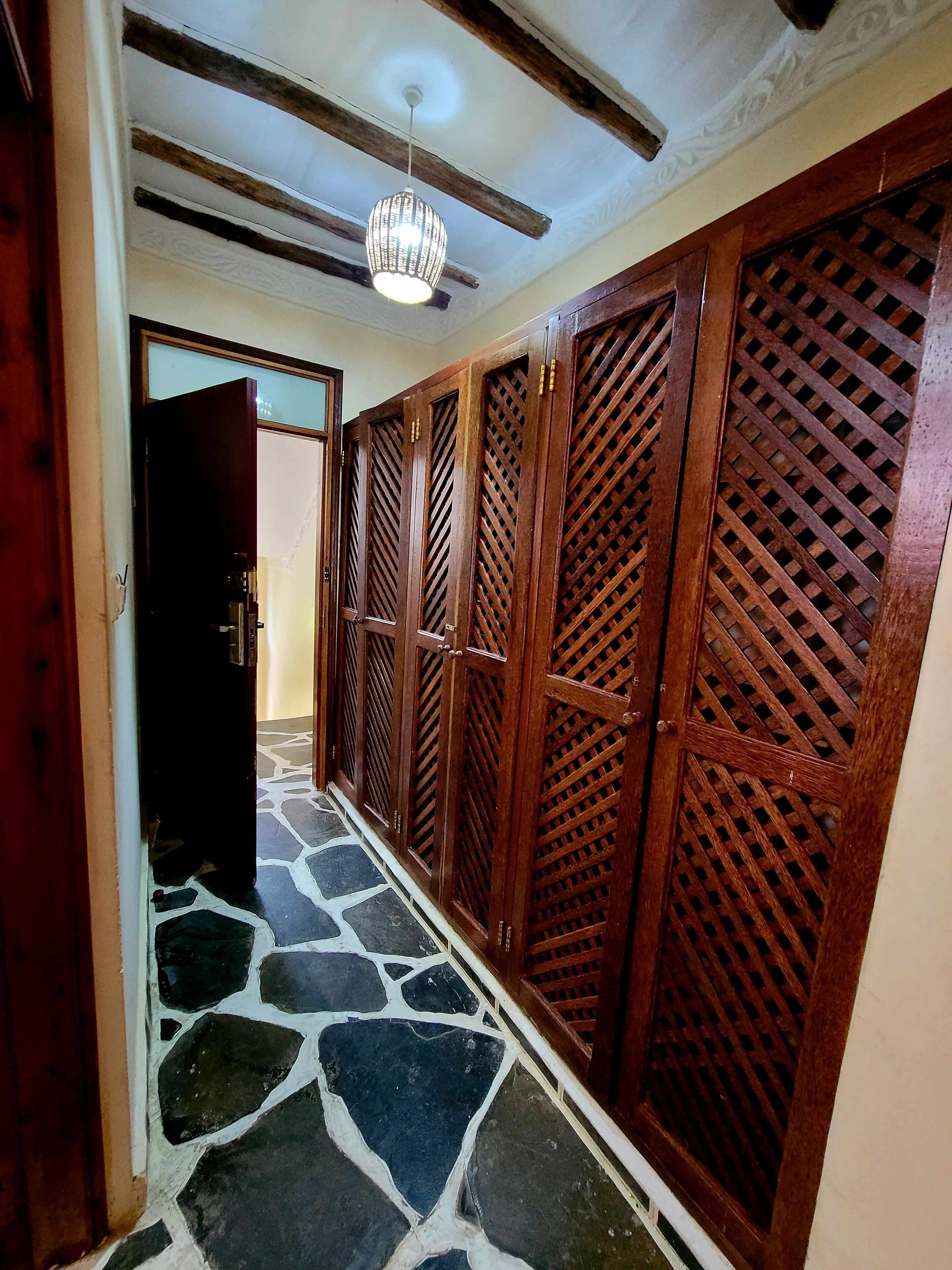 Um corredor com piso de pedra estampada, portas de madeira com desenhos em treliça e um ventilador de teto.
