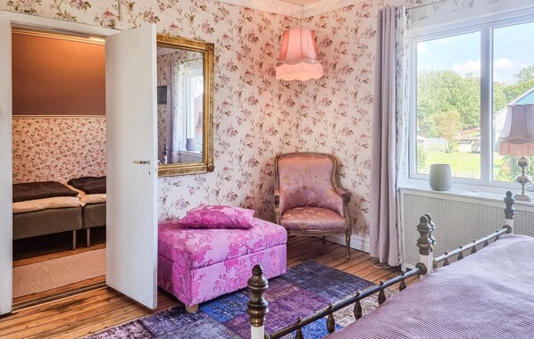 A bedroom with floral wallpaper, a pink ottoman, a chair, a bed, and a window with a view of greenery.