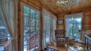 Un acogedor interior de cabaña con ventanas amplias, un sillón estampado con motivos florales, una mecedora y techo de madera.