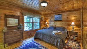 Un dormitorio de cabaña de madera con cama, cómoda, dos mesitas de noche con lámparas, una ventana con cortinas e imagen enmarcada en la pared.