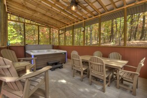 A covered outdoor seating area with wooden chairs and a table, a hot tub, and a view of a forest. - Campbell's Cozy Cabin | Fire Pit | Hot Tub | Grill (Turtletown)