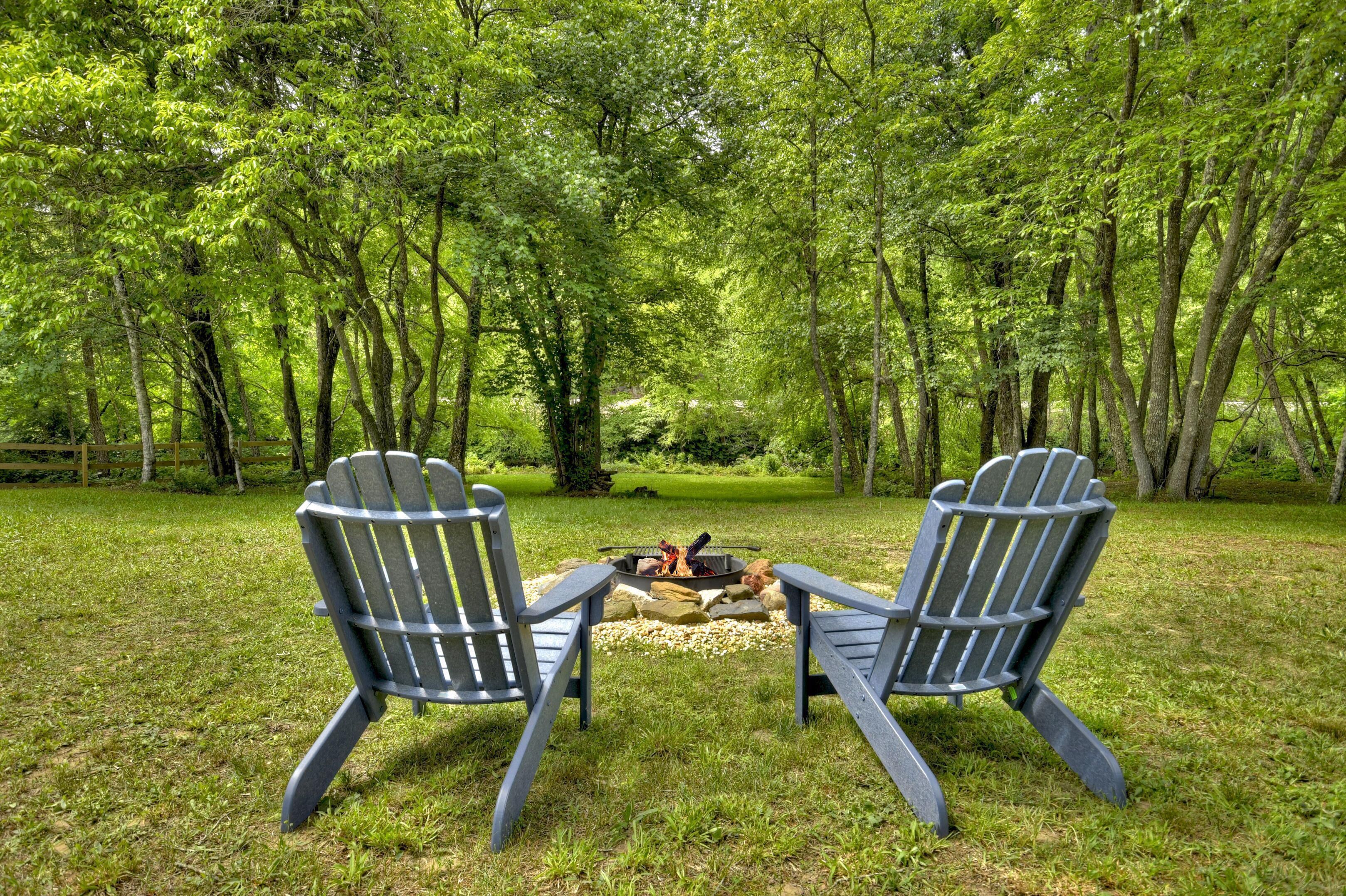 Two Adirondack chairs facing a fire pit surrounded by a lush green forest.