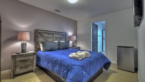 A bedroom with a bed, two bedside tables, a lamp, a refrigerator, and a door leading to a bathroom.