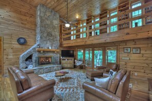 A spacious living room with a stone fireplace, leather sofas, and a large TV. - At Water's Edge | Hot Tub | Fish From Cabin | Secluded (Mineral Bluff)
