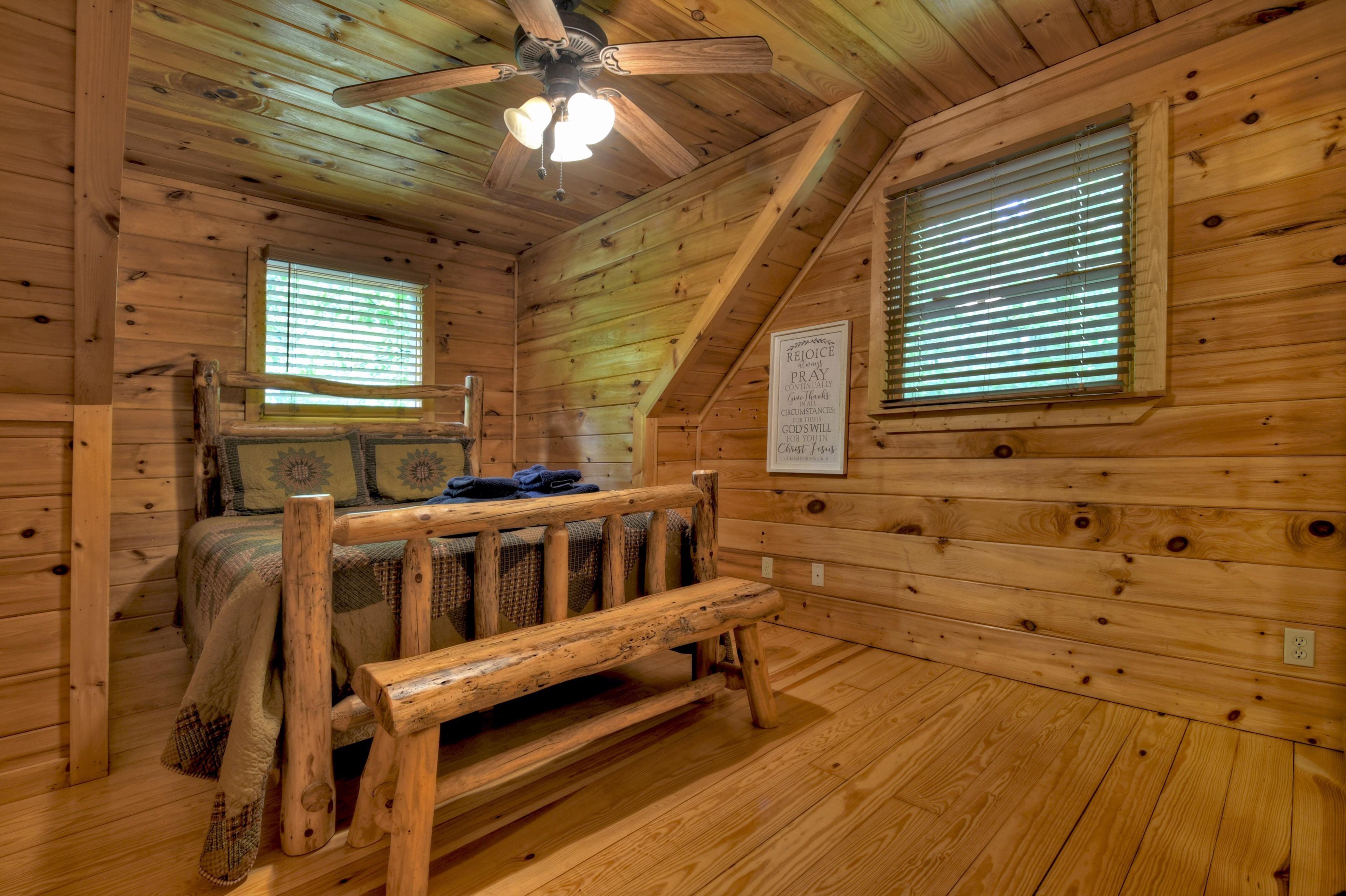 A wooden cabin room with a bed, a bench, a ceiling fan, and two windows with blinds.