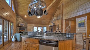 A wooden cabin interior with a kitchen area featuring granite countertops, a dishwasher, and a stone fireplace. - Stokes Haven | Pet Friendly | Hot Tub | Creek (McCaysville)