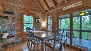 Ein Holzblockhaus-Interieur mit Esstisch, Stühlen, einem Steinkamin und großen Fenstern mit Blick auf den Wald.