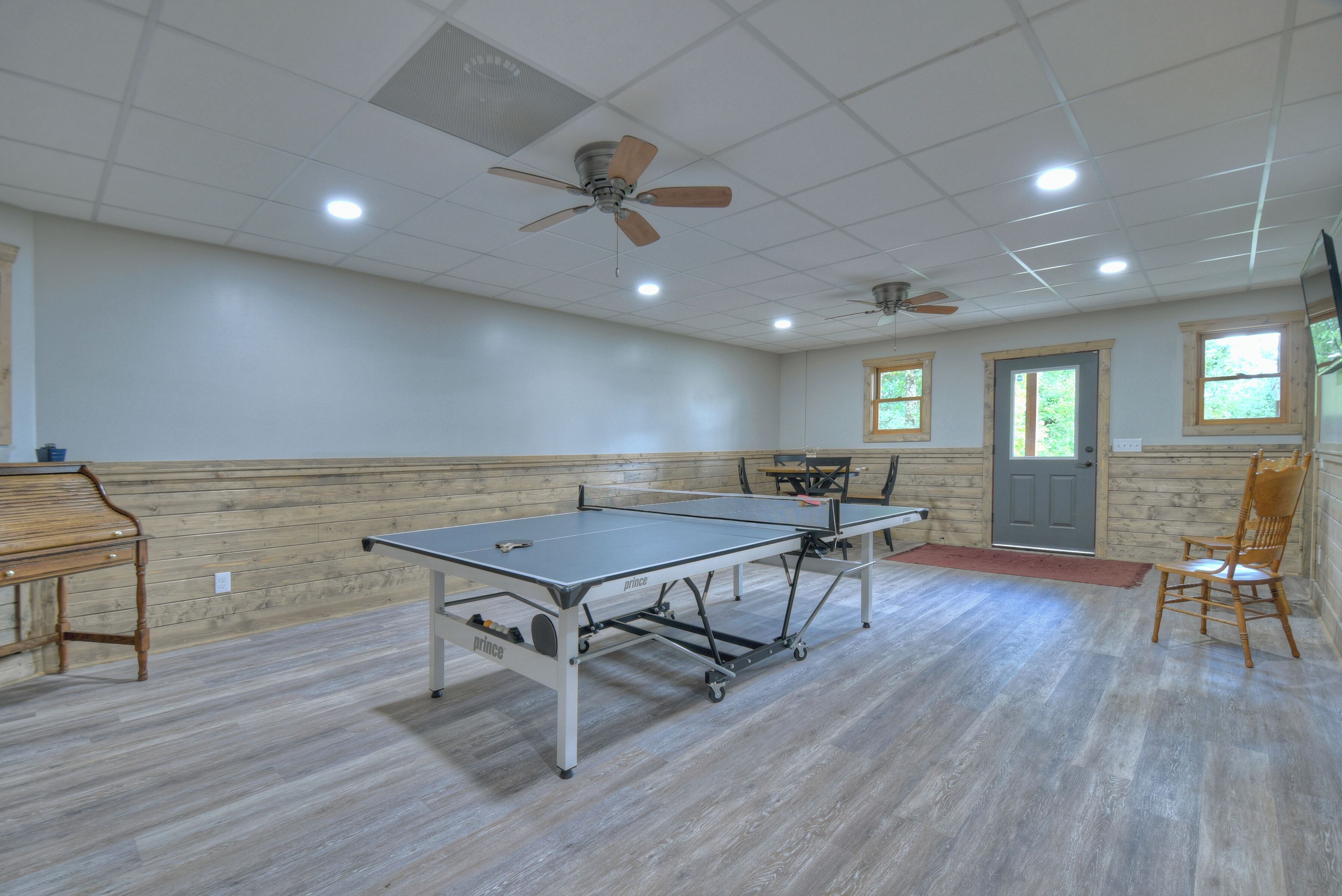 Une pièce spacieuse équipée d’une table de ping-pong, de deux ventilateurs de plafond et d’un piano en bois.