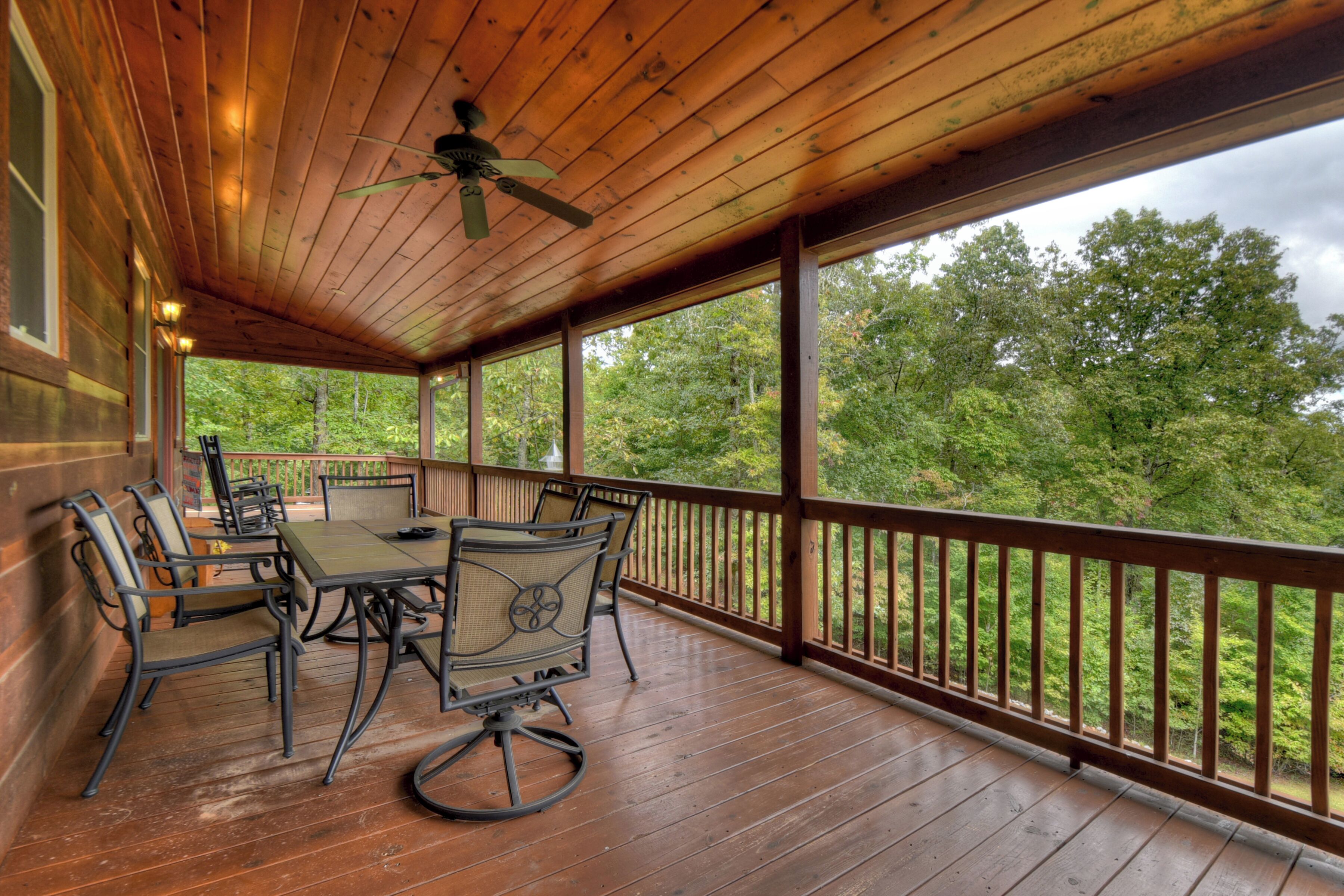 A covered outdoor deck with a wooden table and chairs, a ceiling fan, and a view of a forested area.