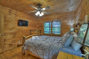 A wooden cabin bedroom with a bed, a ceiling fan, a television, and a window with blinds.