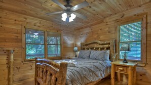Ein Schlafzimmer einer Holzhütte mit Bett, Nachttisch, Deckenventilator und Fenstern mit Jalousien.