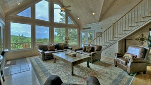 A spacious living room with a wooden ceiling, large windows, a staircase, and leather furniture. - Champagne Chalet | MTN Views | Hot Tub | Secluded (Morganton)