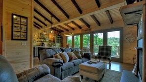 Interior acolhedor de cabana com lareira, área de refeições e uma grande janela com vista para o verde.