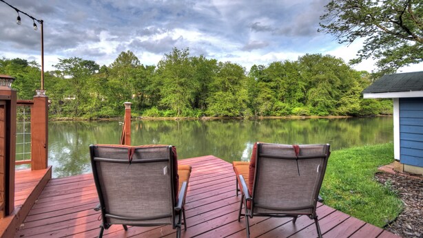 Een houten terras met twee stoelen gericht op een rustig meer, omringd door weelderige begroeiing en een gedeeltelijk bewolkte lucht.