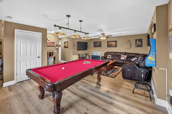 Une table de billard, un canapé et un ventilateur de plafond dans une pièce au plancher en bois et aux murs de couleur beige.