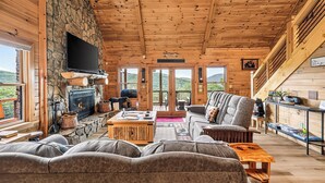 A log cabin interior with a stone fireplace, a flat-screen TV, a sofa, and a dining area. - Shepherd's Sky View | Secluded | MTN Views | Pool Table | (Morganton)