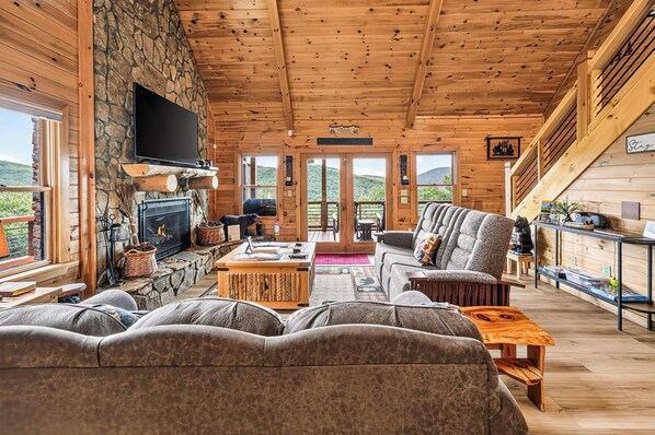 A log cabin interior with a stone fireplace, a flat-screen TV, a sofa, and a dining area.