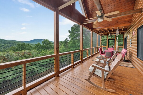 Une terrasse en bois offrant une vue sur un paysage boisé, avec un ventilateur de plafond et une table entourée de chaises.