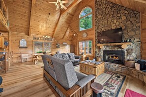 A log cabin interior with a stone fireplace, wooden furniture, and a large TV mounted above it.