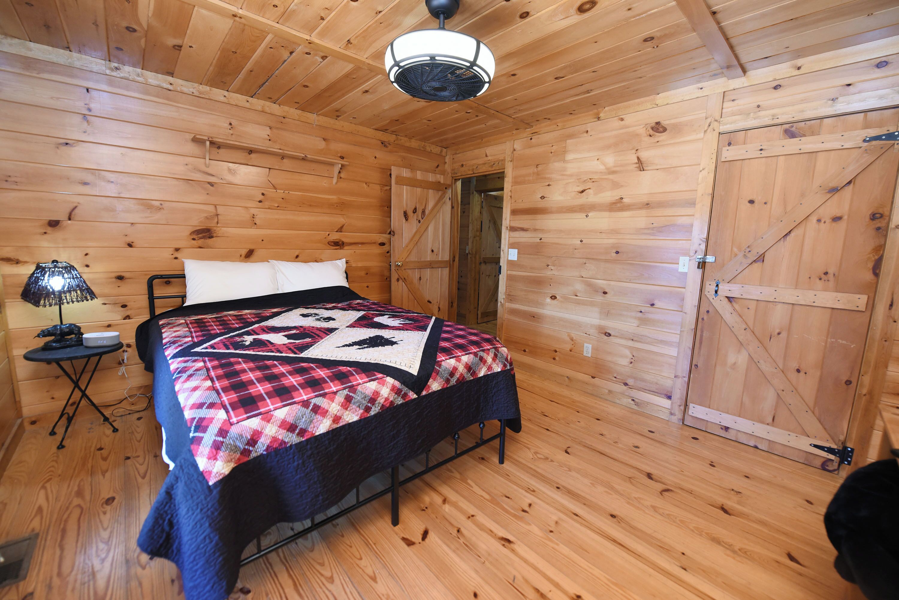 A wooden cabin bedroom with a bed, a small table with a lamp, and a door leading to another room.