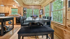 A wooden cabin interior with a dining table, chairs, a built-in oven, and large windows offering a forest view.